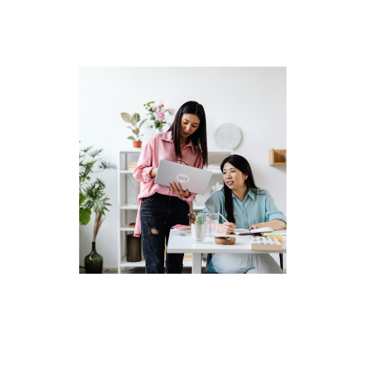 Two women working together in a bright home office. One woman stands beside a desk holding an open laptop and showing the screen, while the other sits at the desk with a notebook and pen, looking at the laptop. Plants and shelves with decor are visible in the background.