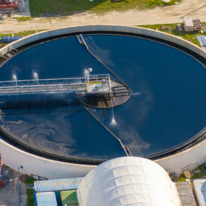 Aerial view of a large circular water treatment tank filled with dark blue water. A central rotating arm sprays water as it moves across the surface, creating ripples and swirling patterns. Surrounding the tank are industrial buildings, pipes, and access roads.