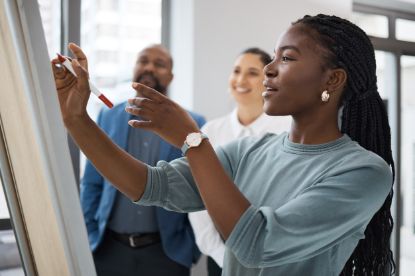 A woman with braided hair writes on a flip chart with a red marker while two colleagues stand behind her, smiling and watching in a bright office setting.