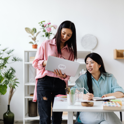 Two women collaborating at a desk in a bright office, one standing and showing something on a laptop while the other sits and writes in a notebook.