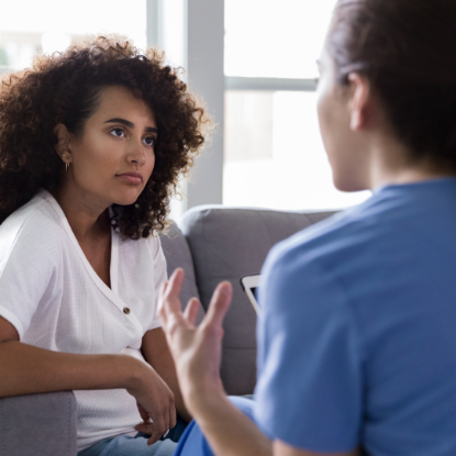 A woman with curly hair sits on a couch, listening attentively while another person, seen from behind, gestures as they speak—suggesting a counseling or serious conversation in a bright room.