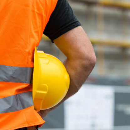 Close-up of a construction worker wearing an orange high-visibility vest and holding a yellow hard hat under their arm at a construction site.