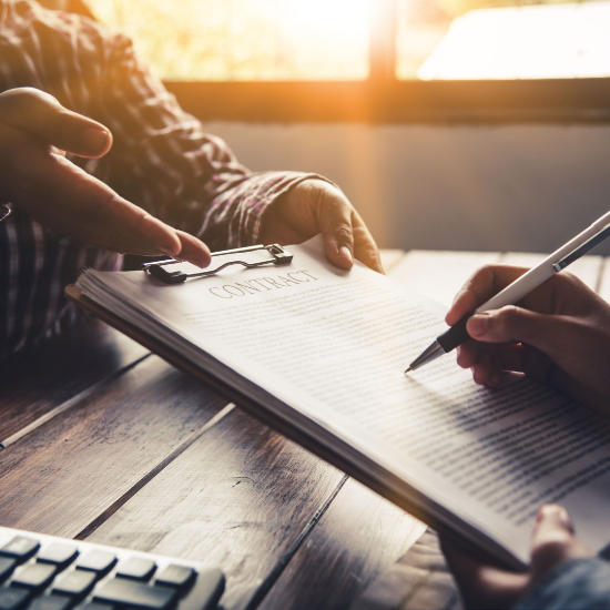Two people reviewing and signing a contract on a clipboard at a wooden table, with sunlight coming through a window and a calculator nearby.