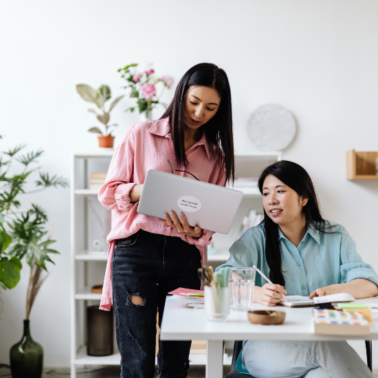 Two women collaborating at a desk in a bright office, one standing and showing something on a laptop while the other sits and writes in a notebook.