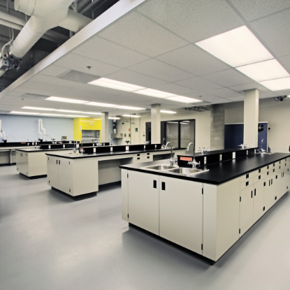 Spacious modern laboratory interior with multiple black-topped workbenches, built-in sinks, storage cabinets, bright overhead lighting, and exposed ventilation ducts on the ceiling.