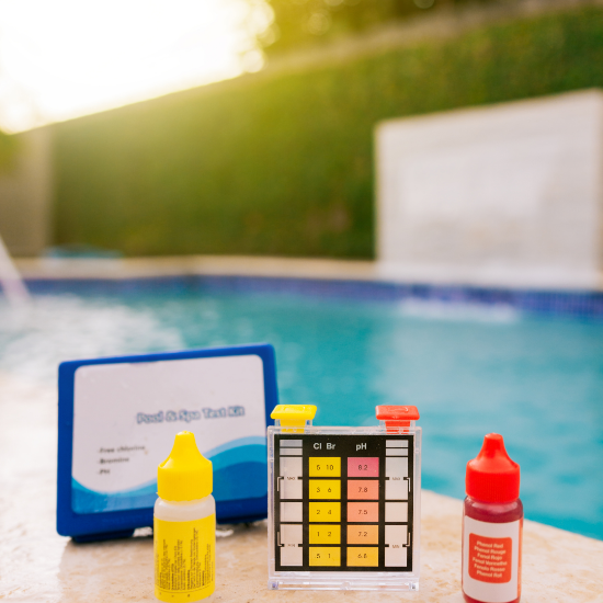 A pool water testing kit placed beside a swimming pool, including a clear comparator block showing chlorine/bromine and pH color scales, a yellow reagent bottle, a red reagent bottle, and a blue case in the background, with the pool water blurred behind.