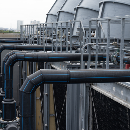Industrial cooling system with large metal pipes, railings, and cylindrical cooling units arranged on a rooftop, with a city skyline faintly visible in the background.