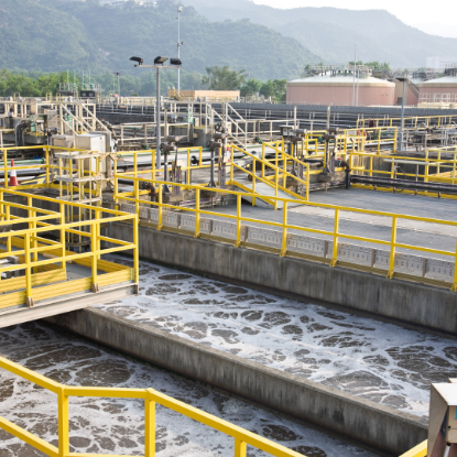 Industrial wastewater treatment facility with concrete aeration basins filled with foamy water, surrounded by yellow safety railings, metal walkways, pipes, and mechanical equipment, with hills in the background.