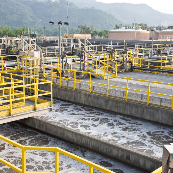 Industrial wastewater treatment facility with concrete aeration basins filled with foamy water, surrounded by yellow safety railings, metal walkways, pipes, and mechanical equipment, with hills in the background.