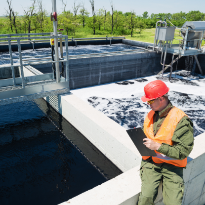 A worker wearing a red hard hat, safety glasses, and an orange reflective vest stands beside a large outdoor wastewater treatment basin, holding a clipboard and inspecting the facility. The basin contains dark water and foam, with pipes, metal walkways, and treatment equipment visible around the tank, while trees and greenery appear in the background.