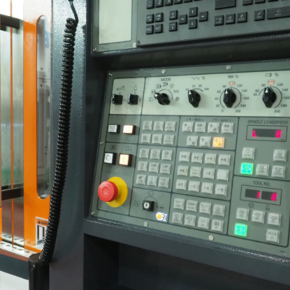 Close-up of a CNC machine control panel with numerous buttons, dials, and a digital display. The panel includes an emergency stop button, keypad controls for machine axes, and illuminated indicators, with part of the machine’s orange safety door and window visible to the left.