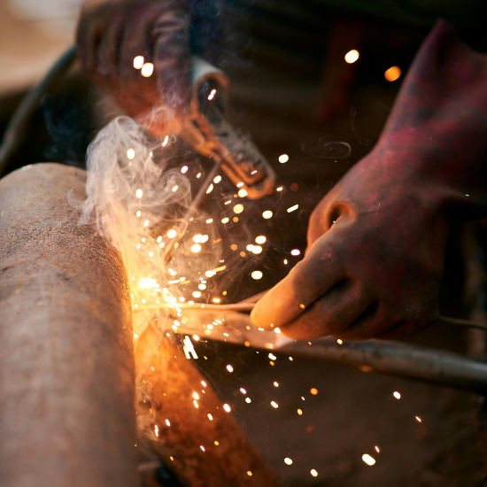 Close-up of a person welding metal with a torch, bright sparks and smoke flying as gloved hands hold the tool against a curved metal surface.