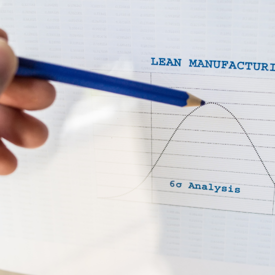 Close-up of a hand holding a blue pencil and pointing to a bell curve chart labeled “6σ Analysis,” with the heading “Lean Manufacturing” visible above, indicating data analysis or process improvement.