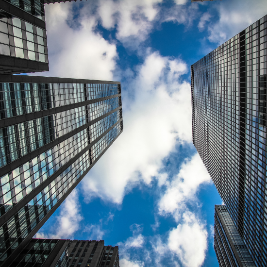 Upward view from street level of several tall glass-and-steel skyscrapers framing a bright blue sky with scattered white clouds.