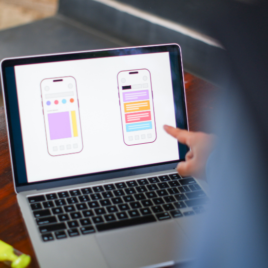 Person pointing at a laptop screen displaying two mobile app interface mockups with colorful layout elements, suggesting UI/UX design or app development.